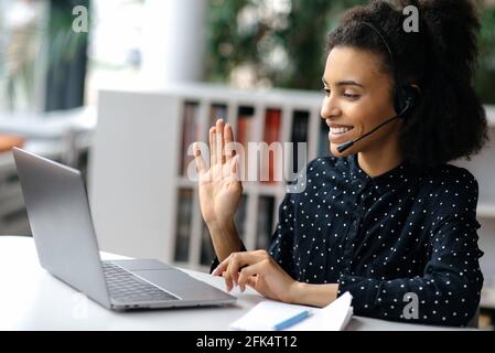 Jeune femme afro-américaine joyeuse et charmante avec micro-casque, responsable ou opérateur de centre d'appels, assise au bureau, ordinateur portable, conversation vidéo avec un client ou un employé, main levée Banque D'Images