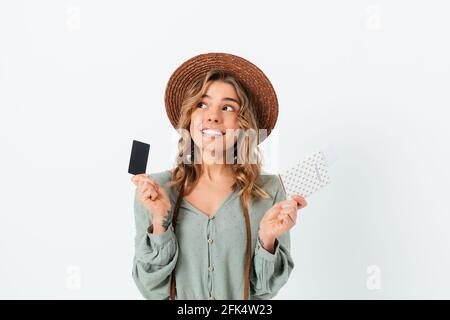 Rêver de vacances jeune femme blonde portant un chapeau de paille d'été carte de crédit et passeport avec billets à la recherche de côté et sourire Banque D'Images