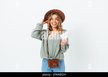 Jolie jeune femme blonde portant un chapeau d'été tenant un passeport avec des billets d'avion, souriant regardant la caméra se tordre, debout sur un studio blanc Banque D'Images