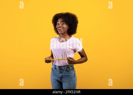 Concept de perte de poids. Ajuster une femme afro-américaine mesurant sa taille avec du ruban adhésif sur fond jaune de studio Banque D'Images