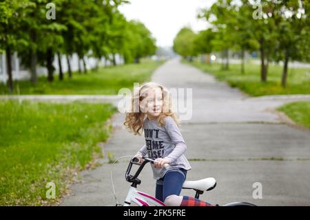 Une petite fille fait du vélo le jour d'été ensoleillé dans le parc de la ville. Les enfants apprennent à conduire un vélo sur une allée à l'extérieur Banque D'Images