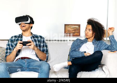 Le couple marié est assis sur le canapé, un gars excité utilise un smartphone et des lunettes VR, joue des jeux virtuels, la fille afro-américaine s'ennuie et elle le regarde de manière indigne Banque D'Images