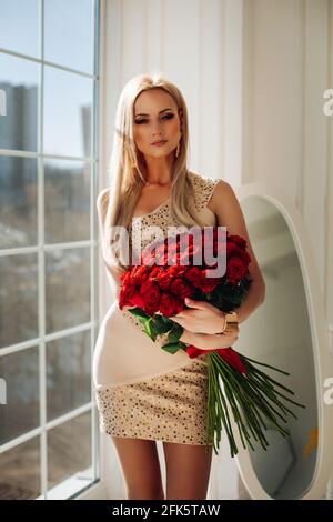 Portrait d'un magnifique modèle blanc blanc en robe beige mini avec des éléments étincelants tenant bouquet de roses rouges debout la fenêtre au soleil Banque D'Images