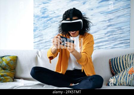 Cyber World, lunettes vr. Joyeuse joyeuse gaie et curieuse jeune afro-américaine assise sur un canapé portant des vêtements décontractés, utilisant un smartphone et testant un appareil de réalité virtuelle dans le monde numérique, souriant Banque D'Images