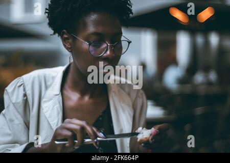 Portrait d'une jeune belle femme noire en lunettes et tranchée blanche, et avec des ongles, assis dans un restaurant le soir et s'étalant Banque D'Images