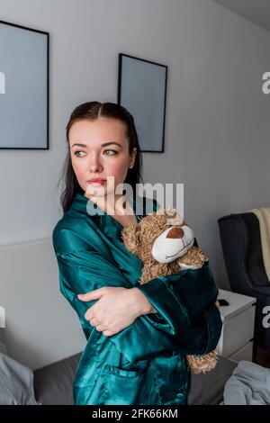 Une jeune femme en pyjama de soie est assise sur le lit et épouse un ours en peluche. Retour aux racines. Concept bien-être. Banque D'Images