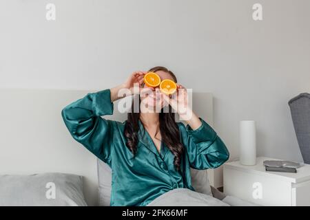 La jeune femme rit et tient les oranges près de ses yeux. Concept de bien-être et zone de confort à la maison. Banque D'Images