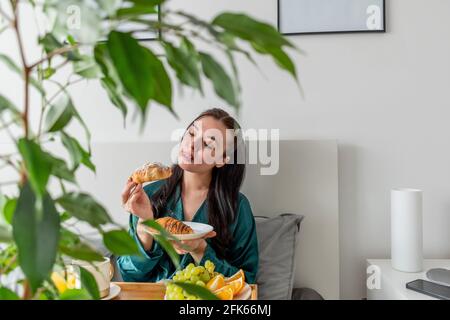 Jeune femme en pyjama de soie a un petit déjeuner sain tout en étant allongé sur le lit dans la chambre. Concept de bien-être à la maison. Santé émotionnelle d'une jeune femme Banque D'Images