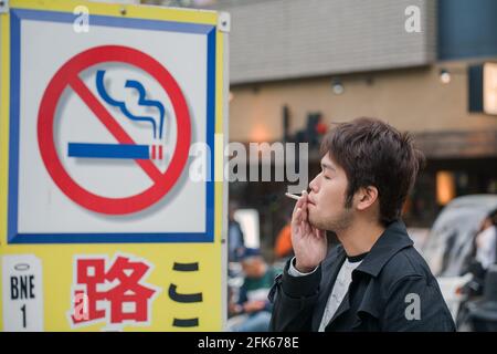 Un homme rebelle japonais se tient à fumer une cigarette à côté d'un panneau 'No Smoking', Akihabara, Tokyo, Japon Banque D'Images