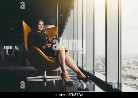 Portrait d'une femme d'affaires caucasienne de taille plus charmante assise un fauteuil orange avec une tablette numérique dans ses mains et en regardant à l'extérieur Banque D'Images