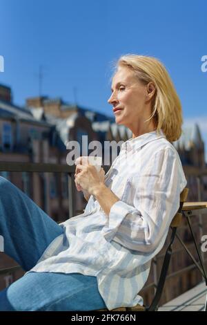 Le matin. Belle femme adulte rêveuse des années 60 dans une tenue décontractée se détendre sur le balcon à la maison, commencer la journée avec le café. Bonne femme âgée assise sur la terrasse et regardant la rue ensoleillée Banque D'Images