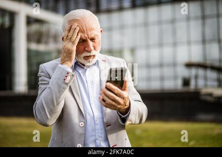 Un homme d'affaires senior est un message sur un smartphone devant le bâtiment de l'entreprise. Il est inquiet. Banque D'Images