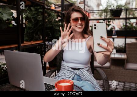 Jeune femme joyeuse utilisant un smartphone pour les appels vidéo pendant qu'elle est assise Dans un café à l'extérieur Banque D'Images