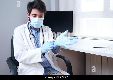 Portrait d'un médecin professionnel et confiant, jeune homme, sous un masque médical et un manteau blanc, stéthoscope au-dessus du cou, portant des gants pendant une pandémie de coronavirus Banque D'Images