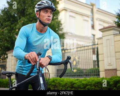 Jeune cycliste masculin portant des vêtements de sport et un casque de protection qui a l'air concentré, debout avec son vélo de route dans le parc pendant la journée Banque D'Images