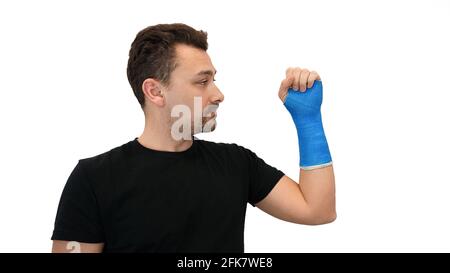 Poignet cassé dans un bandage bleu moderne étanche. Beau barbu d'âge moyen dans un T-shirt noir regarde sa main poing enveloppée dans un plâtre moulé sur un fond blanc Banque D'Images