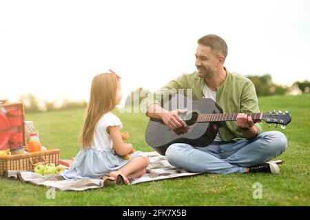 Jolie petite fille dans des vêtements décontractés assis sur un green herbe dans le parc et regarder son papa aimant jouer guitare Banque D'Images