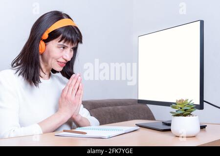 Une femme d'affaires souriante dans un casque regarde un moniteur avec un écran blanc et montre un signe de remerciement, de gratitude, de paumes jointes ensemble, côté Banque D'Images