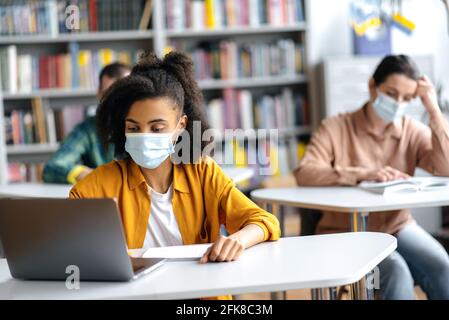 Les jeunes étudiants étudient en cas de pandémie, assis à l'université à distance les uns des autres et portant des masques médicaux protecteurs. Une étudiante afro-américaine prend des notes pendant la conférence, à l'aide d'un ordinateur portable Banque D'Images