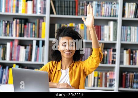 Excellente étudiante féminine. Une jeune fille étudiante afro-américaine intelligente à une conférence est assise à une table, élégamment habillée, lève la main, connaît la réponse à la question et a une idée, veut répondre, sourit Banque D'Images