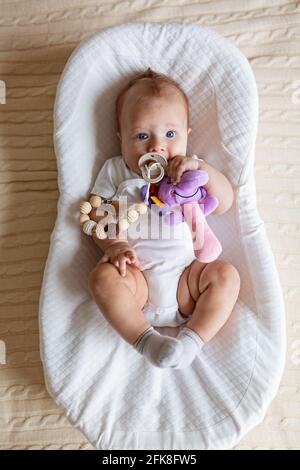 Mignon bébé blond de trois mois couché dans un cocon à la maison. Enfant tient un jouet sonore bourré, suce le mamelon. Banque D'Images