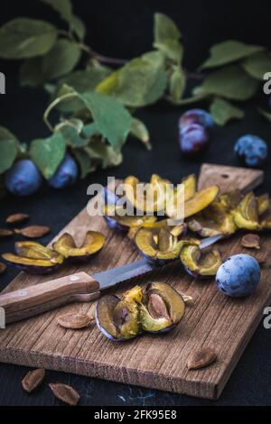 Prunes: prunes mûres en tranches et fruits entiers sur une planche en bois, fond foncé, photo verticale Banque D'Images