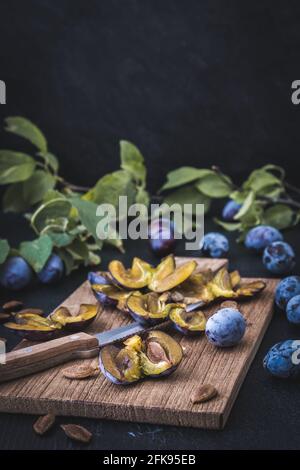 Prunes : prunes mûres en tranches et fruits entiers sur un panneau en bois, fond foncé, photo verticale avec espace de copie Banque D'Images