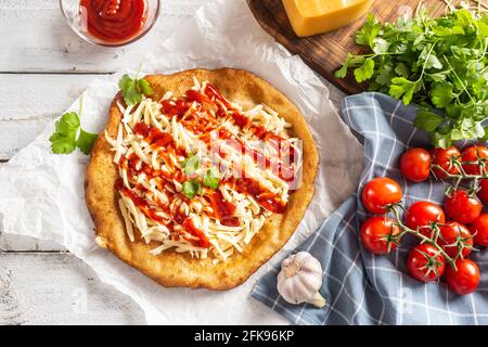 De savoureux langos frits servis sur du papier à pâtisserie avec beaucoup de fromage râpé, ketchup, herbes fraîches et tomates sur un plat à carreaux. Banque D'Images