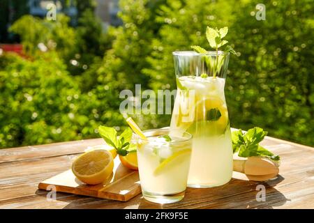 Pichet de limonade glacé, centrifugeuse et un verre de boisson froide aux agrumes avec tranches de citron, feuilles de menthe et pailles jaunes sur une table en bois bruni, avec Banque D'Images