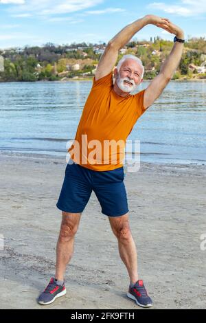 Heureux énergique senior retraité homme s'étendant sur la plage, faisant l'exercice de virage latéral au bord de la rivière. Gros plan. Banque D'Images