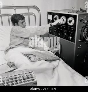 Années 1960, historique, un jeune homme allongé dans un lit d'hôpital en ajustant un cadran sur une grande machine à laquelle il est attaché, une machine de dialectique qui aide à pomper le sang dans les reins, sud-est de Londres, Angleterre, Royaume-Uni. La dialyse est un traitement médical qui filtre et nettoie le sang à l'aide d'une machine, ce qui aide à maintenir l'équilibre du corps lorsque les reins ne peuvent pas exécuter cette fonction. Également connu sous le nom de machines d'hémodialyse. Les machines HD nettoient le sang en le passant à travers un filtre appelé un dialyseur. Banque D'Images