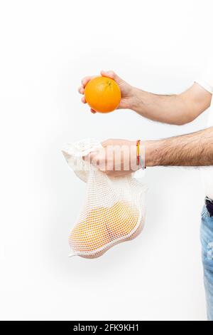 Un homme méconnaissable mettant des oranges dans un sac en filet sur un fond blanc. Copier l'espace. Banque D'Images