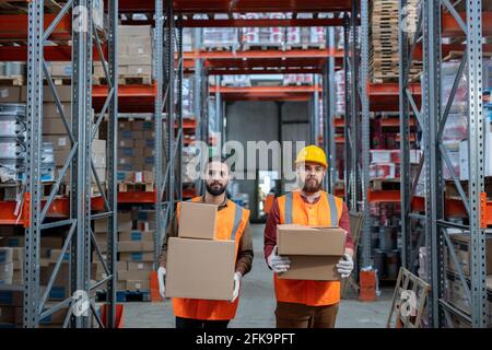 Portrait de grands déménageurs d'entrepôt dans des gilets orange debout avec des piles de boîtes dans l'allée entre les tablettes de cadre en métal Banque D'Images