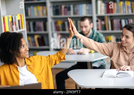 Heureux jeunes étudiants intelligents de différentes nationalités assis à des tables dans la bibliothèque du campus se préparant pour les leçons ou l'examen, deux filles sympathiques joyeuses se donnant cinq autres, souriant Banque D'Images