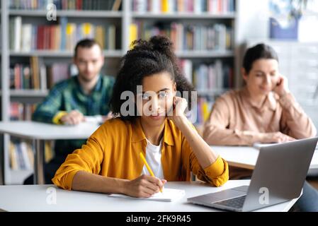 Étudiants en conférence. Concentrée sérieuse afro-américaine étudiante assise à table dans la bibliothèque de l'université, se préparant pour les leçons ou l'examen, regarde la caméra, les étudiants défocused sur le fond Banque D'Images