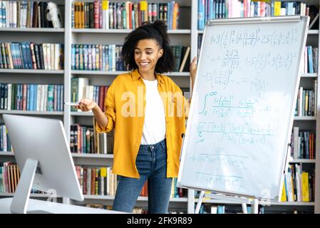 Happy SMART confiante belle enseignante afro-américaine élégante, se tient près du tableau blanc, mène une leçon en ligne, communique avec les élèves par le biais d'une vidéoconférence, par ordinateur Banque D'Images