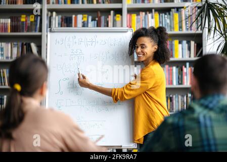 Jeune enseignante afro-américaine à poil dur, agréable et réussie, debout près du tableau blanc de la bibliothèque, donnant une conférence aux étudiants, montrant des informations et souriant Banque D'Images