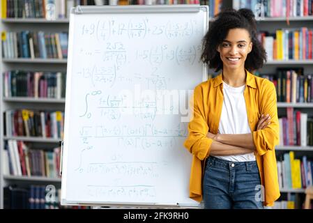 Portrait de mignonne joyeuse fille, enseignante afro-américaine, se tient sur tableau blanc, vêtu de vêtements décontractés, conduit une leçon, croisant ses bras, regardant l'appareil photo, souriant amical Banque D'Images