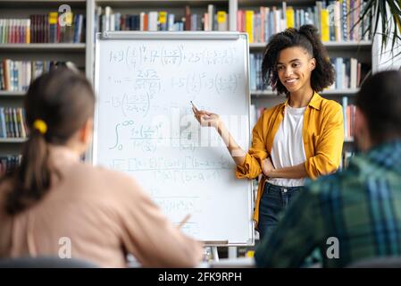 Jeune enseignante afro-américaine à poil dur, agréable et réussie, debout près du tableau blanc de la bibliothèque, donnant une conférence aux étudiants, montrant des informations et souriant Banque D'Images