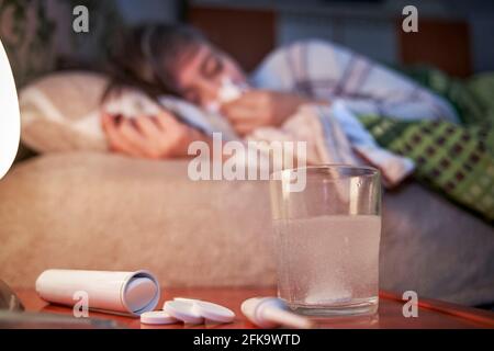 Hors foyer une femme malade couché sur le lit, soufflant son nez avec un tissu. Sur l'accent, sur la table de chevet il y a des médicaments, un verre avec un efferve Banque D'Images