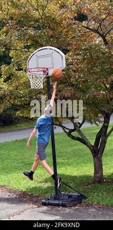 Un jeune adolescent pratique le basket-ball à l'extérieur dans son allée. Il prend un layette et la balle est dirigée vers le panier. Il saute haut. Banque D'Images