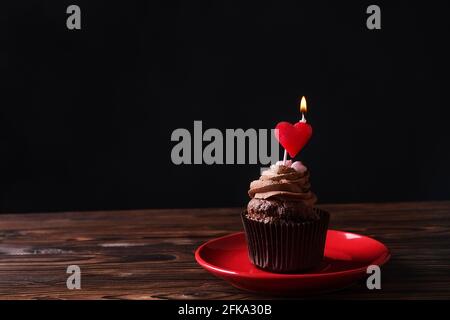 Délicieux cupcakes au chocolat noir, garniture de bougie en forme de coeur brûlant, table texturée en bois. Boulangerie au cacao, café rustique en céramique rouge Banque D'Images