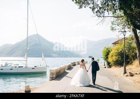 La mariée et le marié marchent main dans la main sur la route au bord de la mer, un yacht blanc est debout près de la rive Banque D'Images