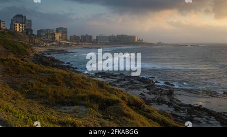 Moody Sunrise se profile pour la ville de Newcastle au-dessus de l'océan. Les vagues du matin se roulent le long du rivage rocheux tandis que les nuages s'amassent au-dessus de la ligne d'horizon Banque D'Images