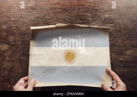 Le drapeau national blanc et bleu de l'Argentine sur un morceau de papier déchiré. Les mains d'un homme pressaient une feuille de parchemin dépliée sur une table en bois marron. VI Banque D'Images