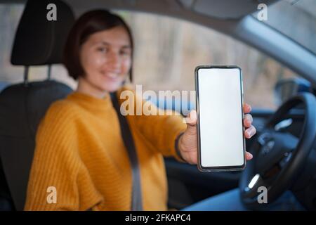 Une jeune femme heureuse conduisant une voiture, elle montre un écran de téléphone vide . Applications mobiles utiles pour le conducteur. Vue latérale à l'intérieur de la voiture, maquette Banque D'Images