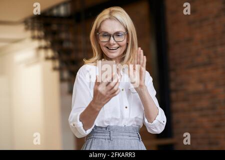 Heureux de À plus. Belle femme mûre avec des cheveux blonds et des lunettes souriant et agitant tout en faisant un appel vidéo à l'aide de son smartphone, debout à Banque D'Images