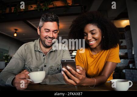 Couple de courses mixtes qui rigole sur des photos sur des réseaux sociaux smartphone dans un café branché Banque D'Images