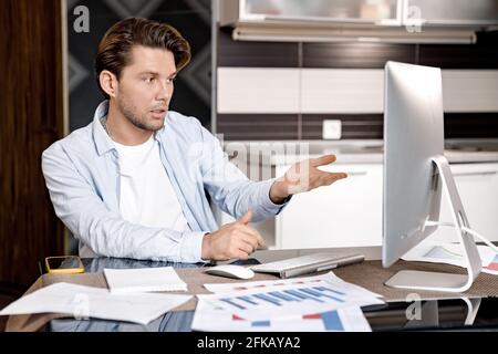 Un homme sérieux est assis au bureau à domicile, en utilisant un ordinateur, en vidéoconférence. Un jeune homme d'affaires qui parle avec lui Banque D'Images