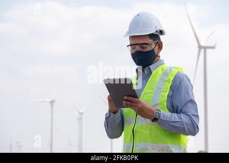 Les moulins à vent d'ingénieur portant un masque facial et un comprimé de honging avec le vent turbine en arrière-plan Banque D'Images